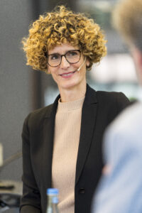 Foto von Mirjam Stegherr während einer Moderation, lächelnd in schwarzem Jacket mit hellem Pullover, zudem trägt sie ein Headset und eine Brille. Die Haare sind sehr lockig, mittelblond und etwa kinnlang. 